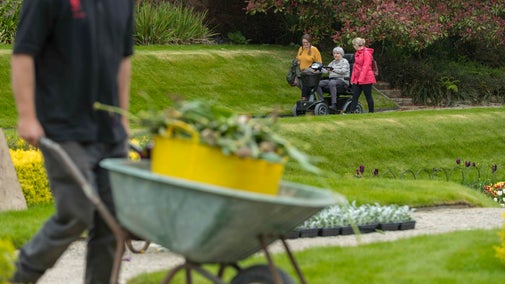 Gardener Pushing Wheelbarrow in sunken garden, with visitors in background.